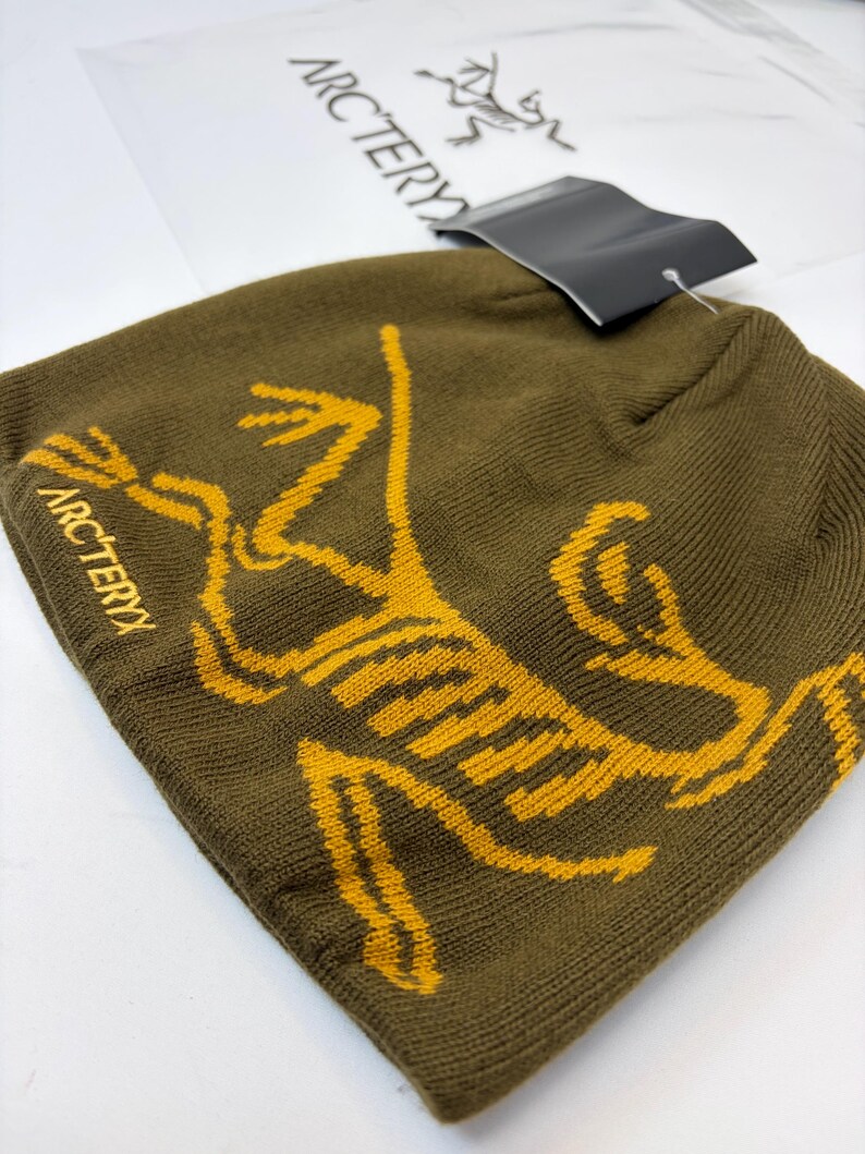 K&ouml;nnte beinhalten: Olivgr&uuml;ne M&uuml;tze mit gelber Grafik eines stilisierten Wesens und dem Wort "ARCTERYX" in Gelb. Ein schwarzes Etikett ist angebracht. Die M&uuml;tze ist aus Strickmaterial.