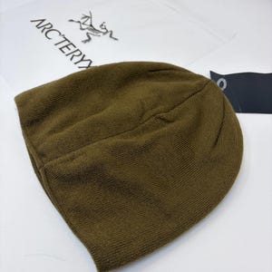 K&ouml;nnte beinhalten: Olivgr&uuml;ne Strickm&uuml;tze. Der Rand ist umgeschlagen. Ein Etikett ist angebracht. Im Hintergrund befindet sich eine transparente Plastikt&uuml;te mit dem Arc'teryx-Logo.