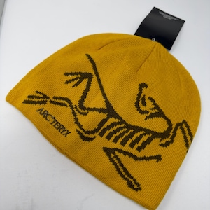 May include: Mustard yellow beanie with a brown graphic of a fossilized bird and the word "ARC'TERYX". The beanie has a tag attached. This knit hat is a winter accessory.