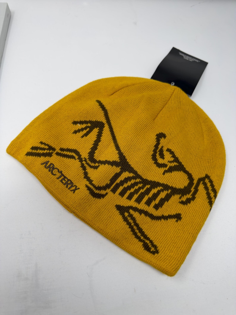 May include: Mustard yellow beanie with a brown graphic of a fossilized bird and the word "ARC'TERYX". The beanie has a tag attached. This knit hat is a winter accessory.