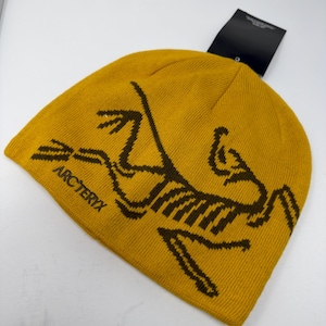 May include: Mustard yellow knit beanie with the brand name "Arc'teryx" and a brown logo. The beanie has a folded brim and a black tag attached. The hat is made of a soft, warm material.