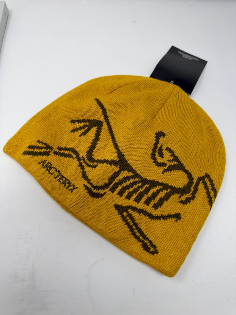 May include: Mustard yellow knit beanie with the brand name "Arc'teryx" and a brown logo. The beanie has a folded brim and a black tag attached. The hat is made of a soft, warm material.