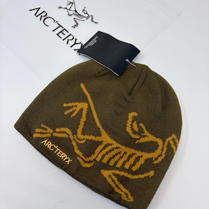 K&ouml;nnte beinhalten: Olivgr&uuml;ne M&uuml;tze mit gelbem Aufdruck und dem Text "ARC'TERYX". Die M&uuml;tze ist aus Strickmaterial und hat ein Etikett. Eine transparente Plastikt&uuml;te mit dem Markenlogo befindet sich im Hintergrund.
