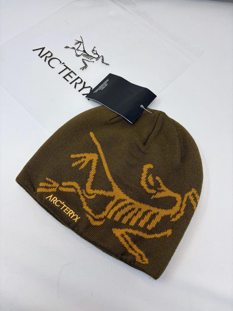 K&ouml;nnte beinhalten: Olivgr&uuml;ne M&uuml;tze mit gelbem Aufdruck und dem Text "ARC'TERYX". Die M&uuml;tze ist aus Strickmaterial und hat ein Etikett. Eine transparente Plastikt&uuml;te mit dem Markenlogo befindet sich im Hintergrund.