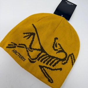 May include: Mustard yellow beanie with a brown graphic design and the word "ARC'TERYX" printed on the side. The beanie has a tag attached. This winter hat is made of a soft, knitted material.