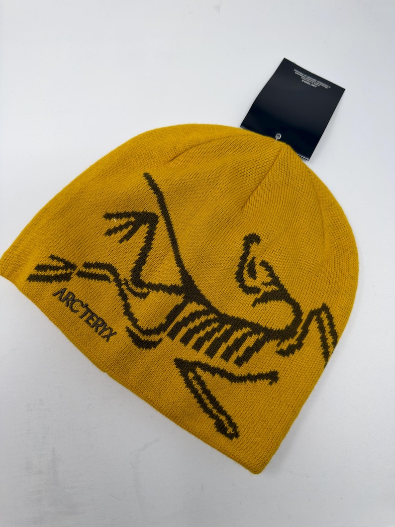 May include: Mustard yellow beanie with a brown graphic design and the word "ARC'TERYX" printed on the side. The beanie has a tag attached. This winter hat is made of a soft, knitted material.