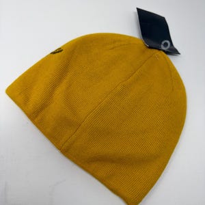 May include: Mustard yellow knit beanie hat with a small logo on the side. The hat has a ribbed texture and a black tag attached. The beanie is made of a soft, warm material, perfect for cold weather.