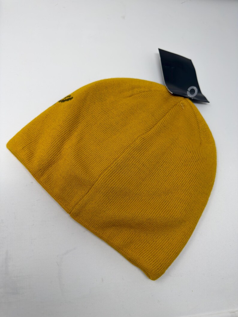 May include: Mustard yellow knit beanie hat with a small logo on the side. The hat has a ribbed texture and a black tag attached. The beanie is made of a soft, warm material, perfect for cold weather.