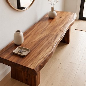 Può includere: Una lunga panca in legno con bordo naturale, caratterizzata da una ricca venatura del legno marrone e due robuste gambe a blocco. Sulla superficie si trova un piccolo vassoio in ceramica beige contenente chiavi in metallo e un vaso in ceramica.