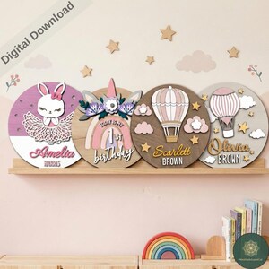 Può includere: Quattro decorazioni murali circolari in legno con nomi personalizzati. I disegni includono un coniglietto, un unicorno, mongolfiere e il testo "Today is my 1st birthday". La tavolozza dei colori comprende rosa, bianco, marrone e grigio.