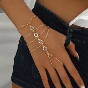 May include: Gold-toned hand chain bracelet with clear, round gemstones. The bracelet features multiple delicate chains that connect to a ring worn on the middle finger. The jewelry is worn on a wrist and hand.