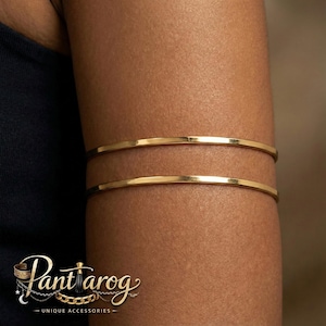 May include: Two gold-colored arm cuffs are worn on the upper arm. The jewelry is simple and elegant, with a smooth, polished finish. The cuffs are thin and circular, and they appear to be made of metal. The logo "Pantarog - Unique Accessories" is visible in the lower left corner.
