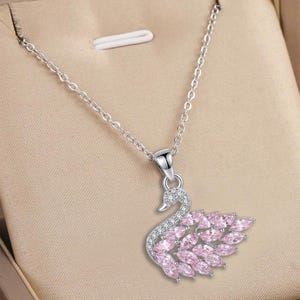 Può includere: Una collana color argento con un ciondolo a forma di cigno. Il collo e la testa del cigno sono incastonati con piccole pietre chiare, mentre l'ala è composta da diverse pietre rosa chiaro taglio marquise su una montatura in metallo argentato.