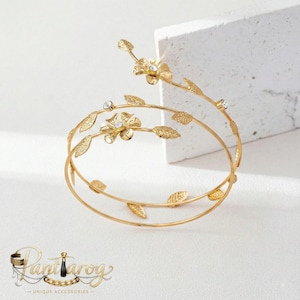 May include: Gold-toned arm cuff with a floral and leaf design. The cuff features two flowers with crystal accents and several leaves. The cuff wraps around the arm with a spiral design. The jewelry is on a white background.