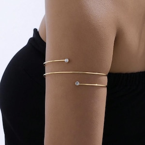 May include: Gold-toned arm cuff jewelry with two delicate bands. Each band features a small, clear gemstone. The jewelry is worn on the upper arm, creating a minimalist and elegant look. The background is a dark top.