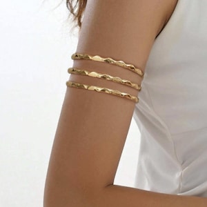 May include: Gold-toned arm cuffs. The image shows three wavy, textured arm cuffs worn on the upper arm. The jewelry has a shiny finish and is displayed against a neutral background. The cuffs are a simple yet elegant accessory.