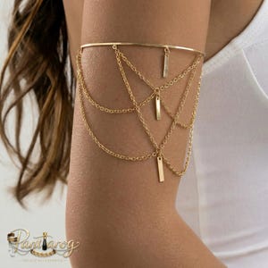 May include: Gold-toned arm cuff featuring a delicate design of chains and rectangular bar accents. The upper arm jewelry has a simple band that connects to multiple chains that drape down the arm. The arm cuff is worn on the upper arm.