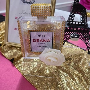 May include: A clear glass bottle with a gold lid and a label that reads "No. 16 Deana Paris Parfum". The bottle is sitting on a gold sequined surface with a white rose.