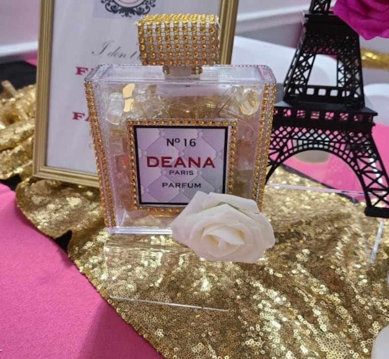 May include: A clear glass bottle with a gold lid and a label that reads "No. 16 Deana Paris Parfum". The bottle is sitting on a gold sequined surface with a white rose.