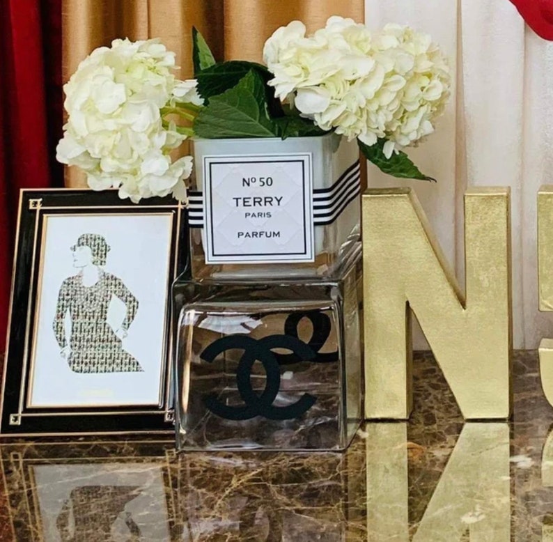 May include: A clear glass vase with white hydrangeas sits on a table with a gold letter N and a framed black and white illustration of a woman. The vase has a label that reads "N&deg;50 TERRY PARIS PARFUM".
