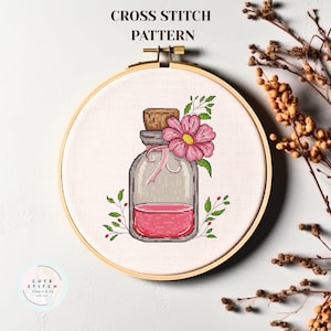 Può includere: Schema a punto croce con una bottiglia di vetro con un tappo di sughero, riempita di liquido rosa. Un fiore rosa e foglie verdi adornano la bottiglia. Il disegno è in un telaio da ricamo in legno. Il testo in alto recita "CROSS STITCH PATTERN".