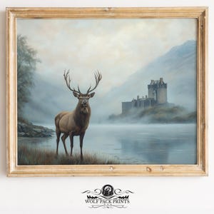 Puede incluir: Una impresión enmarcada de un paisaje con un majestuoso ciervo junto a un lago, con un castillo al fondo. La obra de arte tiene una estética vintage, con colores suaves y un marco de madera. La impresión es de Wolf Pack Prints.