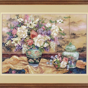 Puede incluir: Pintura enmarcada de naturaleza muerta floral. Un gran jarrón de flores blancas, rosas y moradas se encuentra sobre una mesa con un paño dorado. Un cuenco más pequeño de flores y un jarrón decorativo también están sobre la mesa. El fondo presenta un paisaje.