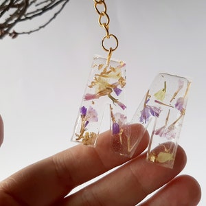 Puede incluir: Un llavero de cadena dorada con una letra 'M' de resina transparente incrustada con flores secas y escamas doradas.