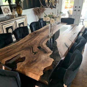 Puede incluir: Una gran mesa de comedor rectangular con una tapa de madera de borde vivo y un río de resina epoxi negra. La mesa está rodeada de sillas tapizadas negras. La veta de la madera es una mezcla de tonos marrones claros y oscuros.