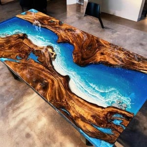 May include: A rectangular wooden table with a blue epoxy resin river design. The table features natural wood grain in shades of brown and tan, contrasting with the blue and white resin that resembles ocean waves. The table is supported by black legs.