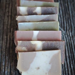 以下が含まれることがあります： 茶色、白、ピンク、緑など、さまざまな色と模様の8つの手作り石鹸の積み重ね。石鹸は木製の表面に垂直に並べてあります。
