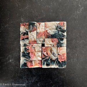 May include: A collection of rectangular handmade soaps, each with a unique swirl design. The soaps feature shades of pink, white, grey, and teal. They are arranged on a dark, textured surface.