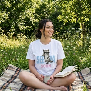 Può includere: T-shirt bianca con grafica di procione e la scritta "SEE YA SOON". La maglietta è indossata all'aperto su una coperta a quadri. Una persona tiene in mano una tazza e un libro.