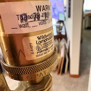 Puede incluir: Primer plano de un portal&aacute;mparas de lat&oacute;n con una etiqueta que dice "WARN" y "Wildwood Lampholder". La etiqueta tambi&eacute;n incluye "E-51077" y "UL PORTABLE LAMP ISSUE NO. BG-508". El portal&aacute;mparas tiene un anillo texturizado y estampado.