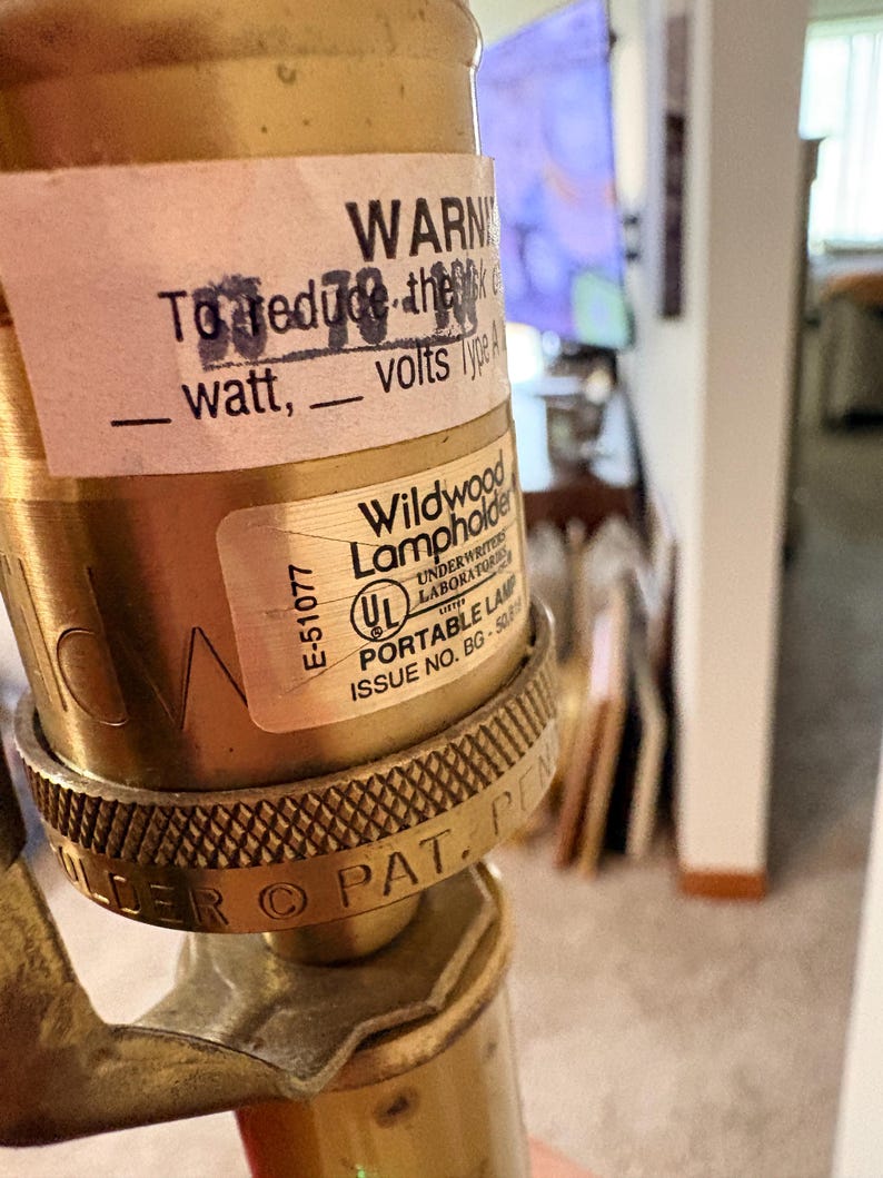 Puede incluir: Primer plano de un portal&aacute;mparas de lat&oacute;n con una etiqueta que dice "WARN" y "Wildwood Lampholder". La etiqueta tambi&eacute;n incluye "E-51077" y "UL PORTABLE LAMP ISSUE NO. BG-508". El portal&aacute;mparas tiene un anillo texturizado y estampado.