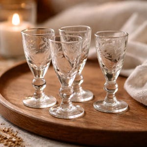 Puede incluir: Cuatro copas de licor de vidrio transparente grabadas con un diseño floral, exhibidas en una bandeja redonda de madera. Las copas tienen una base con tallo y un borde acampanado. Una vela encendida es visible en el fondo.