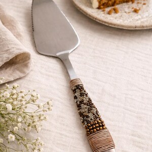 May include: A stainless steel cake server with a beaded handle. The handle features a mix of brown, gold, and clear beads, wrapped with a copper-colored wire. A slice of cake with white frosting sits on a plate in the background.