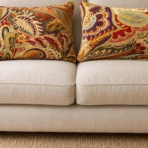 Può includere: Due cuscini rettangolari con un vivace motivo paisley nei toni del rosso intenso, arancione bruciato, verde oliva e oro. I cuscini sono posizionati fianco a fianco su un divano imbottito color beige chiaro con un tessuto liscio e tinta unita.