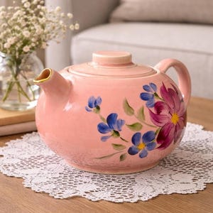 May include: A pink ceramic teapot with a lid and handle, decorated with hand-painted blue and purple flowers. The spout has a gold rim. The teapot sits on a white lace doily.