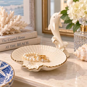 May include: A cream-coloured ceramic trinket dish shaped like a scallop shell with a gold-painted rim. A white ceramic dolphin figurine stands upright on the edge of the dish. Inside the dish are a pearl bracelet and two gold-toned rings.