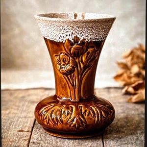 May include: A brown ceramic vase with a flared top and a bulbous base. The vase features a floral relief design and a textured, off-white rim. The vase is approximately 8 inches tall and sits on a wooden surface.