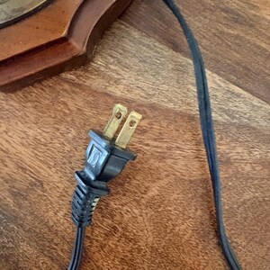 Puede incluir: Primer plano de un enchufe el&eacute;ctrico negro con dos clavijas de lat&oacute;n y un cable negro. El enchufe est&aacute; conectado a un cable negro y el fondo es una superficie de madera. El enchufe es un enchufe el&eacute;ctrico est&aacute;ndar.
