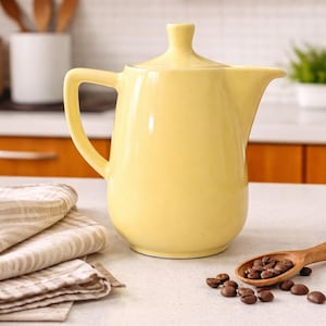 Puede incluir: Una cafetera de cerámica amarillo pálido con tapa, asa y boquilla, colocada sobre una encimera blanca. Una cuchara de madera con granos de café y un paño plegado a rayas beige y blanco están cerca.