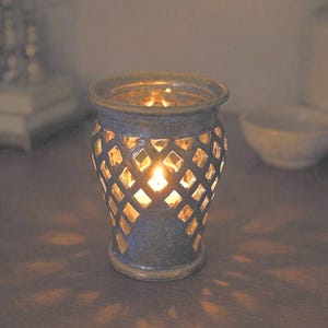 May include: A blue ceramic candle holder with a diamond-patterned design. The candle holder has a cylindrical shape with a flared top and base. A lit candle inside casts a warm glow through the cutouts, creating a cozy ambiance.