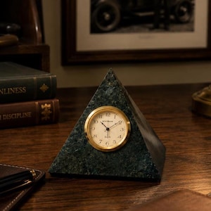 May include: A pyramid-shaped desk clock with a gold-colored clock face and hands. The clock is set in a dark green marble base. The clock is on a wooden desk with books and a pen.