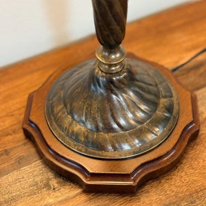 Puede incluir: Primer plano de una base de l&aacute;mpara vintage. La base presenta una columna de metal oscuro y retorcido que se eleva desde una placa de metal circular decorativa. La placa se asienta sobre una base octogonal de madera. La base de la l&aacute;mpara tiene una est&eacute;tica cl&aacute;sica y antigua.