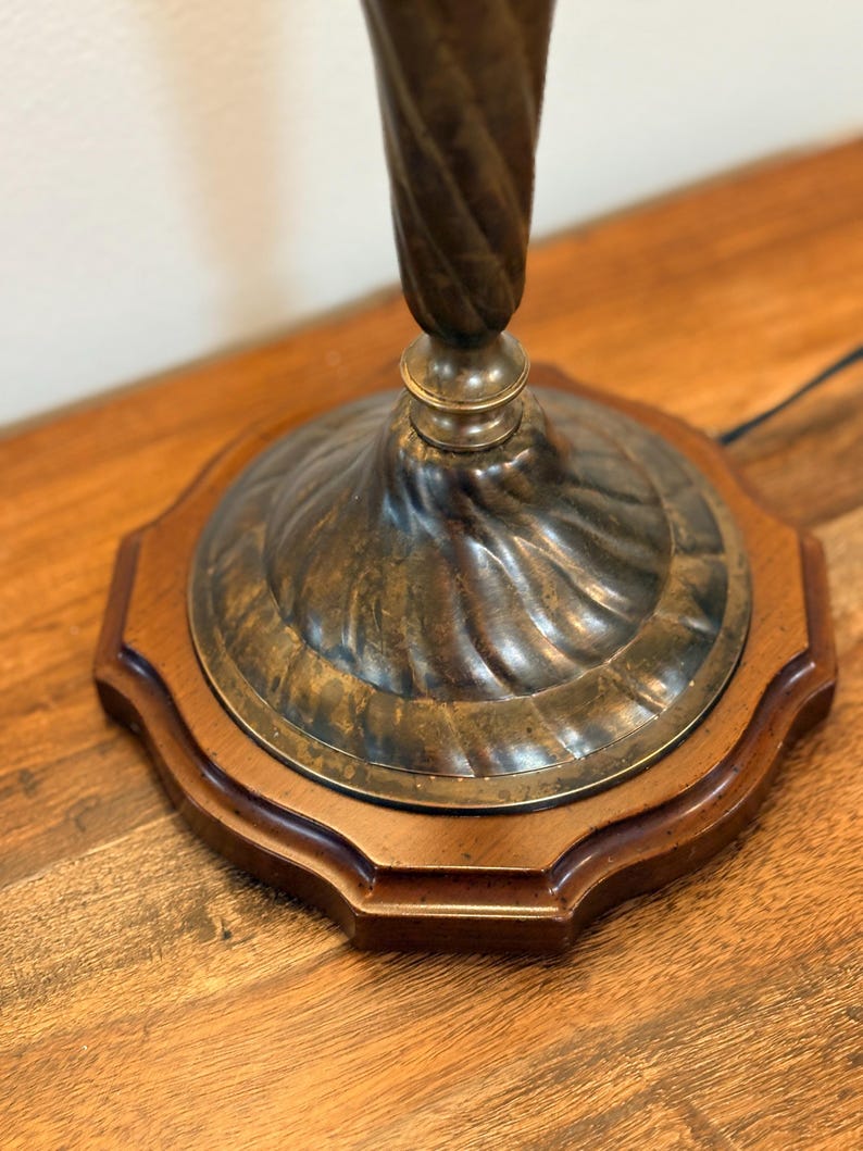Puede incluir: Primer plano de una base de l&aacute;mpara vintage. La base presenta una columna de metal oscuro y retorcido que se eleva desde una placa de metal circular decorativa. La placa se asienta sobre una base octogonal de madera. La base de la l&aacute;mpara tiene una est&eacute;tica cl&aacute;sica y antigua.