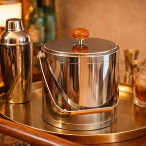 Könnte beinhalten: Ein Edelstahl-Eiskübel mit Holzgriff und Deckel, ein Cocktailshaker und ein Getränk mit einer Orangenscheibe auf einem goldfarbenen Tablett. Die Barutensilien stehen auf einem hölzernen Servierwagen.