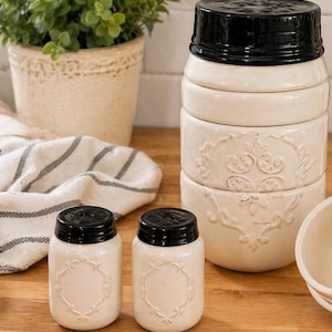 May include: Cream-colored ceramic kitchen canisters and salt and pepper shakers with black lids. The canisters are stacked, and the shakers have a decorative embossed design. A potted plant and a striped towel are in the background.