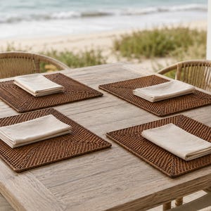 May include: Four square brown woven rattan placemats arranged on a light-colored wooden table. Each placemat features a folded off-white cloth napkin placed in the center. The placemats have a detailed, textured weave pattern with a border.